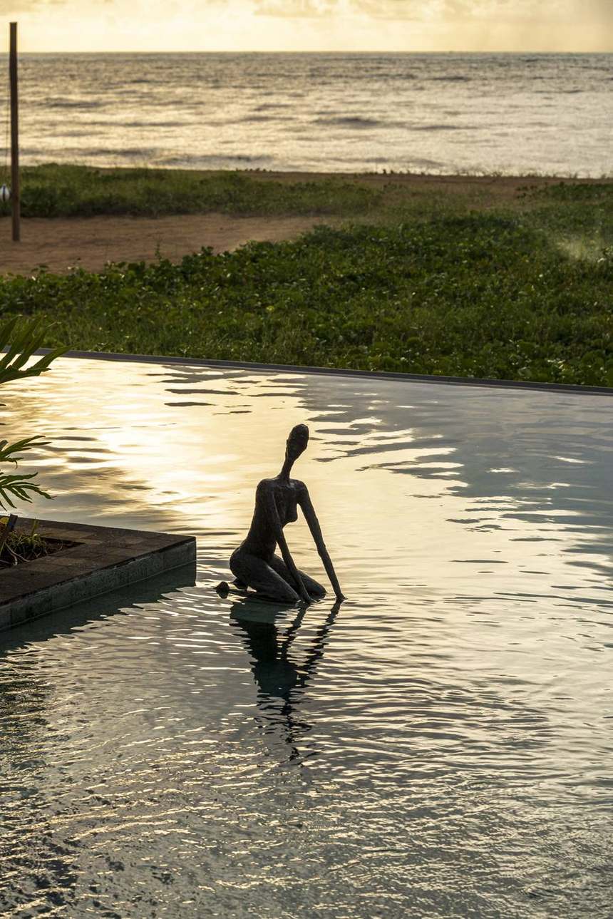 Escultura na borda infinita da piscina ao pôr do sol, com o mar ao fundo